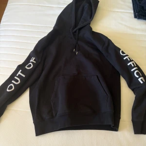 Svart hoodie Wood Wood  - Svart hoodie med huva och känguruficka framtill. På båda ärmarna står det 'OUT OF OFFICE' i stora vita bokstäver. Klassisk passform och dragsko i huvan, perfekt för en chill look.