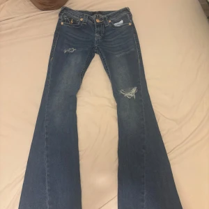 Blå bootcut jeans från True Religion - Säljer ett par blå jeans från True Religion med bootcut passform och slitna detaljer på framsidan. Jeansen har klassiska fickor med dekorativa sömmar och guldknappar både fram och bak. Perfekt för dig som gillar en avslappnad och cool stil.