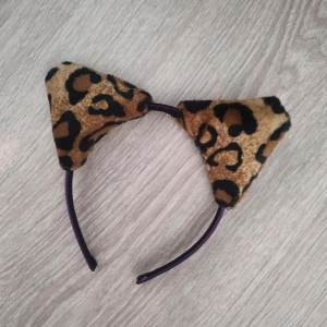 Diadem med leopardmönstrade kattöron - Ett diadem med mjuka, trekantiga kattöron i leopardmönstrat tyg. Öronen är bruna och svarta med en fluffig känsla, perfekt för att sticka ut på maskerad eller temafest. Diademet är svart och smalt, sitter bekvämt på huvudet.