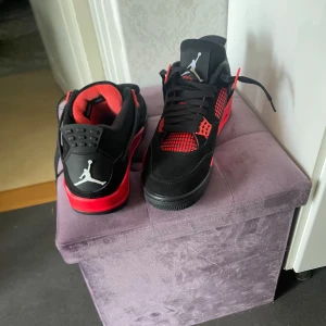 Nike Air Jordan 4 svart/röd sneakers - Säljer ett par Nike Air Jordan 4 sneakers i svart och rött med klassisk Jumpman-logga på hälen och sulan. Skorna har svarta snören, meshdetaljer och röda accenter på sula och snörning. Ovandelen är i mocka och textil, med en riktigt clean och sportig vibe. Storlek 44 men passar även 43 lika bra