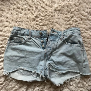Ljusblå jeansshorts med rå kant - Säljer ett par ljusblå jeansshorts från 157 med hög midja och rå, fransig kant. Shortsen har klassisk femficksdesign, synliga knappar framtill och slitna detaljer för en chill vibe. Perfekta för varma dagar och enkel att matcha med din favorit-t-shirt.