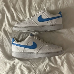 Nike Air Force 1 vit/blå sneakers - Snygga Nike Air Force 1 sneakers i vitt skinn med blå swoosh och detaljer. Klassisk låg modell med perforerad tå, vit sula och snörning. Finns ej kartong till har använt dom 2 gånger