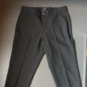 Svarta kostymbyxor från Jack & Jones - Stilrena svarta kostymbyxor från Jack & Jones med klassisk rak passform och hällor för bälte. Perfekta för en clean och dressad look. Byxorna har knapp och dragkedja framtill samt diskreta sidfickor.