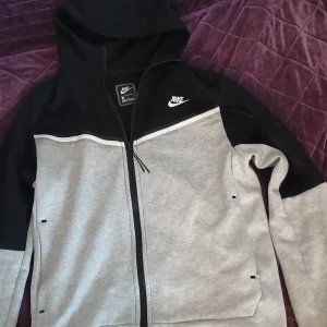Nike hoodie i svart och grå med dragkedja - Nike hoodie i svart och ljusgrått med vit logga på bröstet. Tröjan har hel dragkedja, huva och långa ärmar. Snygg kontrast mellan färgerna och sportig look. Perfekt för chill eller träning. Klassisk streetstyle från Nike.