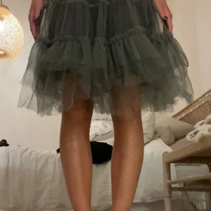 Mörkgrå volangkjol från Holly Bracken - Supersnygg mörkgrå kortkjol från Holly Bracken med flera lager volanger i tunn polyester. Kjolen har en fluffig och trendig look som ger extra volym och är perfekt för dig som vill sticka ut. Elastisk midja för skön passform.