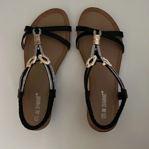 Svarta sandaler med glittriga detaljer - Snygga svarta sandaler från J.M. Diamant med glittriga pärldetaljer och guldfärgade dekorationer på remmarna. Platt sula och öppen tå gör dem perfekta för varma dagar. Stilren design med bruna innersulor och svarta remmar som ger en elegant touch.