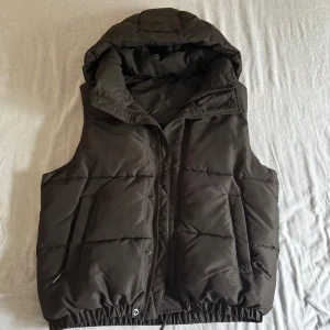 Zara Väst - Snygg svart dunväst från Zara med stor vadderad huva och dragkedja framtill. Västen har två fickor på sidorna och elastisk nederkant för en skön passform. Perfekt att slänga över en hoodie när det är kyligt ute. Inga tecken på slitage, ser helt ny ut