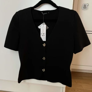 Svart stickad topp från Gina Tricot - Svart stickad topp från Gina Tricot med korta ärmar och v-ringning. Toppen har silvriga knappar framtill i och en figurnära passform. 