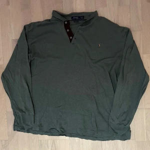Grön långärmad pikétröja Polo Ralph Lauren - Snygg mörkgrön långärmad pikétröja från Polo Ralph Lauren med klassisk krage och bruna knappdetaljer. Tröjan har broderad logga på bröstet och är gjord i mjuk bomull. Perfekt för dig som gillar stilrena och tidlösa plagg.