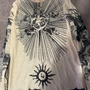 Beige meshtröja med tryck - Säljer en beige meshtröja med långa ärmar och svart tatueringsinspirerat print över hela tröjan. Framsidan har ett stort motiv med texten 'Safe Sex For Ever' och coola detaljer som drakar och sol. Perfekt för dig som vill sticka ut med edgy stil.