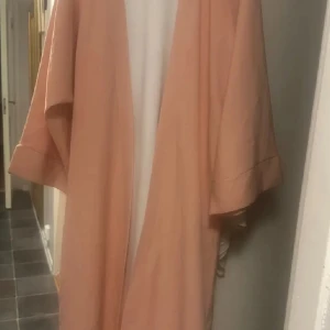 Lång ljusrosa omlottklänning - Säljer en lång, ljusrosa/aprikos färgad  2 delad kläning  med vida ärmar och ledig passform. Klänningen har en enkel och stilren design, perfekt för dig som gillar en flowy och chill vibe. Materialet känns mjukt och lätt mot huden.