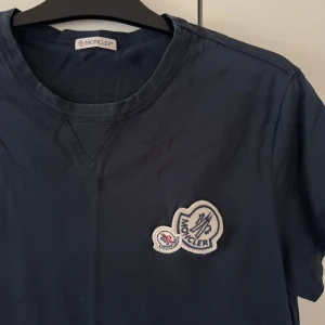 Mörkblå Moncler t-shirt med patch - Snygg mörkblå t-shirt från Moncler med rund hals och korta ärmar. På bröstet sitter två broderade Moncler-patchar i vitt och rött som ger en cool detalj. T-shirten är gjord i mjuk bomull och har en klassisk passform som funkar till allt.