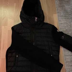 Svart Moncler cardigan med huva - Svart pufferjacka från Moncler med stickade ärmar och vadderad kropp. Jackan har huva med dragsko, dragkedja framtill och Moncler-logga på ärmen. Snygga detaljer som dragkedjeficka på ärmen och ribbade muddar. Perfekt för kalla dagar och riktigt stilren look. Snabb affär=bättre pris!