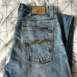 Nudie Jeans blå jeans 32/34 - Snygga ljusblå jeans från Nudie Jeans med klassisk femficksdesign och kontrastsömmar. Jeansen har en rak passform och cool slitning framtill samt Nudie-logga på bakfickan. Perfekta för en avslappnad streetstil.