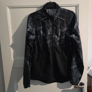 Svart vindjacka SOC med mönster - Svart vindjacka från SOC i storlek S med coolt abstrakt mönster i vitt och grått. Jackan har hel dragkedja, ståkrage och två sidofickor. Materialet är lätt och syntetiskt, perfekt för träning utomhus. Reflekterande detaljer på ryggen bröstet och ärmen för extra synlighet.