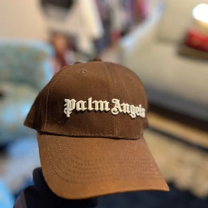 Brun Palm Angels keps  - Snygg brun keps från Palm Angels med broderad vit logga framtill och på baksidan. Kepsen har en klassisk böjd skärm, justerbart metallspänne och en liten palm-broderi på sidan. Storleken är juster bar.
