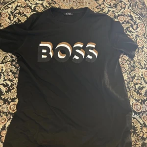 Svart BOSS t-shirt med tryck - Svart t-shirt från Hugo Boss i storlek M. Framsidan har ett stort BOSS-tryck i vitt och brunt med coolt grafiskt mönster. Klassisk rund hals och korta ärmar. Skön bomullskvalitet som känns mjuk mot huden. Perfekt för dig som gillar stilrena logotoplagg. Pris kan minskas