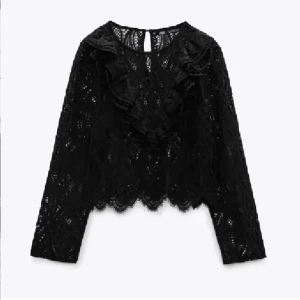 Svart spetsblus med volangdetaljer - Säljer en svart blus i spets med långa ärmar och snygga volangdetaljer framtill. Blusen har ett genombrutet mönster och rund halsringning, samt en croppad passform med vågig kant nedtill. Perfekt för dig som gillar statement-plagg!