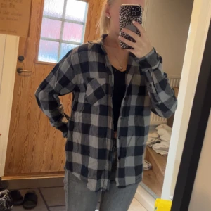 Flanellskjorta - Säljer en oversized rutig flanellskjorta i svart och grått med klassisk krage och knäppning framtill. Skjortan har långa ärmar och en bröstficka. Perfekt att bära öppen över en t-shirt eller stängd för en chill look.