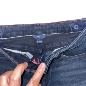Mörkblå jeans från Tommy Hilfiger - Snygga mörkblå jeans från Tommy Hilfiger med klassisk femficksdesign och röda detaljer på insidan av linningen. Jeansen har raka ben och är tillverkade i mjukt denimtyg med lätt tvättad look. Perfekta för en cool och avslappnad stil.