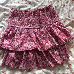 Rosa volangkjol med mönster - Supersöt kort volangkjol i rosa med paisleyinspirerat mönster. Kjolen har smock i midjan för en bekväm passform och är gjord i ett lätt och luftigt polyestertyg. Perfekt för varma dagar när du vill ha något både snyggt och bekvämt. Fungerar som både xs och s