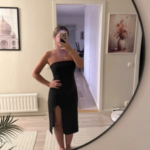 Svart axelbandslös klänning med slits - Snygg svart axelbandslös klänning med slits framtill. Klänningen är figurnära och har en stilren, enkel design som ger en elegant look. Perfekt för dig som vill sticka ut med en klassisk och tidlös stil.