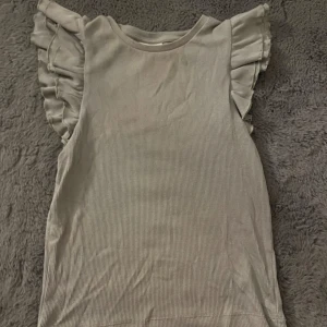 Grön ribbad topp med volangärmar - Supersöt grön topp med ribbad struktur och volangdetaljer på axlarna. Ärmlös modell som ger en luftig och trendig känsla, perfekt att styla med jeans eller kjol. Enkel men ändå extra tack vare volangerna.