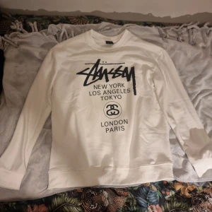 Vit Stüssy sweatshirt med tryck(world tour) - Vit sweatshirt från Stüssy i storlek L med svart tryck både fram och bak. Tröjan har klassisk crewneck, långa ärmar och breda muddar. Materialet är mjuk bomull och passformen är normal. Perfekt för dig som gillar streetwear och vill sticka ut med ikoniskt tryck.