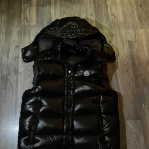 Svart dunväst från Moncler med huva - Säljer en svart, glansig dunväst från Moncler med avtagbar huva och klassisk logga på bröstet. Västen har dragkedja framtill och två fickor. Insidan har Monclers ikoniska serietidningspatch. Perfekt för lager-på-lager och streetstyle.