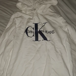 Vit hoodie från Calvin Klein Jeans - Säljer en vit hoodie från Calvin Klein Jeans med stor CK-logga i mörkblått på bröstet. Tröjan har huva och långärmade ärmar, perfekt för chill dagar. Mjuk bomullskänsla och klassisk passform. Snygg och enkel att matcha med jeans eller joggers.