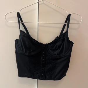 Svart korsettopp  - Snygg svart korsett från gilly hicks/hollister med tunna justerbara axelband och dekorativ spets längs bysten och framtill. Framsidan har hakknäppning. Passar exemeplvis till fest. Använd ett fåtal gånger.