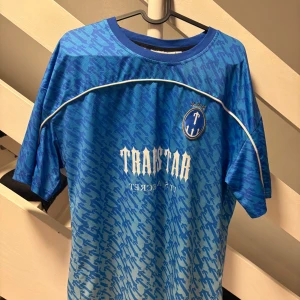 Blå Trapstar fotbollströja S - Snygg blå fotbollströja från Trapstar med vit logga och nummer 22 på ryggen. Tröjan har ett randigt mönster i olika blå nyanser, vit piping och rund hals. Materialet är lätt och ventilerande polyester, perfekt för träning eller match.