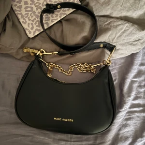 Svart axelväska  - Säljer en svart axelväska med snygga guldfärgade kedjedetaljer och dragkedja upptill. Väskan är i slät skinnimitation och har ett stilrent, rundat formspråk. Perfekt för dig som vill ha en trendig och lyxig accessoar med plats för det viktigaste.