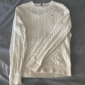 Vit kabelstickad tröja Tommy Hilfiger - Vit kabelstickad tröja från Tommy Hilfiger med klassisk rund halsringning och diskret logga på bröstet. Tröjan har långa ärmar och ribbade muddar. Köptes för runt 900kr, passar runt längden 165. Har andvänts knappast, ungefär 3 gånger. Inga hål, inga fläckar. Perfekt inför hösten och vintern.