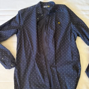 Mörkblå prickig skjorta Lyle & Scott - Snygg mörkblå skjorta från Lyle & Scott med vita prickar och klassisk gul logga på bröstet. Skjortan har lång ärm, knappar framtill och en stilren passform som funkar till många tillfällen.