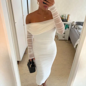 Vit offshoulder långklänning mesh - Supersnygg vit långklänning med offshoulder och långa meshärmar. Klänningen är figurnära med draperad effekt och har en elegant, stilren look. Perfekt för dig som vill sticka ut med en modern och trendig stil.