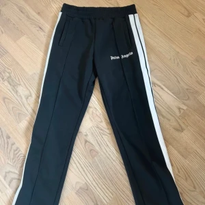 Palm Angels svarta trackpants M - Svarta trackpants från Palm Angels med vita sidoränder och logga tryckt på vänster ben. Byxorna har elastisk midja, raka ben och är gjorda i 100% polyester. Snygg streetstil med sportig vibe och praktiska fickor framtill.