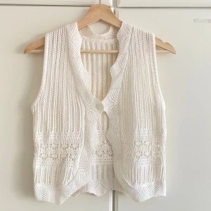 Vit stickad väst med knapp - Superfin vit stickad väst med ett hålmönster och en knapp framtill. Västen har v-ringning och en vågig kant nertill som ger en extra touch. Perfekt att slänga över en t-shirt eller skjorta för en chill och trendig look.