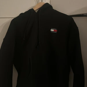 Svart hoodie från Tommy Jeans - Svart hoodie från Tommy Jeans med klassisk logga på bröstet. Tröjan har huva med snörning och en stor magficka. Perfekt för dig som gillar en enkel och stilren look med streetkänsla.