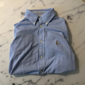 Blå skjorta från Ralph Lauren - Klassisk ljusblå skjorta från Ralph Lauren med broderad logga på bröstet. Skjortan har lång ärm, knappar framtill och button down-krage. Tillverkad i mjuk bomull med Oxfordväv och stilrena vita knappar. Perfekt för en clean och tidlös look.
