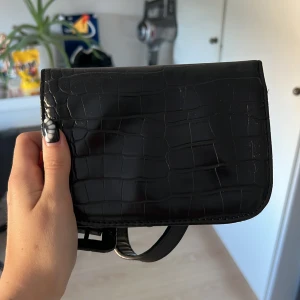 Svart croco väska från BIKBOK - Snygg svart väska från BIKBOK i croco-imitation med justerbart band och fyrkantigt spänne. Väskan har magnetlås och ett huvudfack med dragkedja inuti. Perfekt storlek för det viktigaste och ger en trendig touch till din outfit.