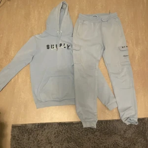 Ljusblå hoodie från Supply - Säljer en ljusblå hoodie från Supply med tryck på bröstet och klassisk känguruficka. Tröjan har huva med snörning och långa ärmar. Perfekt för chill dagar eller när du vill ha en avslappnad stil.