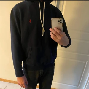 Mörkblå hoodie från Polo Ralph Lauren - Svart hoodie från Polo Ralph Lauren med klassisk röd broderad logga på bröstet. Tröjan har huva med vita snören och en stor magficka. Perfekt för dig som gillar stilren streetwear med premiumkänsla.