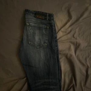 DSQ2 mörkblå jeans med slitningar - Mörkblå jeans från DSQ2 med snygga slitningar och tvättade detaljer. Klassisk femficksmodell med knappgylf och tydlig logotyp på bakfickan. Jeansen har en rak passform och är tillverkade i robust denim för en cool och avslappnad look.