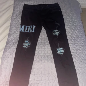 Svarta Amiri jeans med blå detaljer - Säljer ett par svarta jeans från Amiri med blåa tryck och slitningar på benen. Jeansen har slim fit och coola patch-detaljer samt stora bokstäver i blått på låret och bak. Perfekta för dig som vill sticka ut med streetwear-vibe.