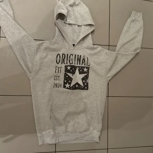 Grå hoodie  - Säljer en ljusgrå hoodie från Original Fit med svartvit stjärntryck och texten 'Original Fit Est. 2024' på bröstet. Tröjan har huva, känguruficka och långa ärmar. Perfekt för dig som gillar en chill och stilren look. Den passar och folk som har XS 