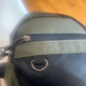 Grön och svart ryggsäck med dragkedja - Ryggsäck i grön och svart färg med praktisk dragkedja framtill och en metallring för extra fästmöjligheter. Tillverkad i slitstark syntet, perfekt för dig som vill ha en enkel och funktionell väska till vardagen.