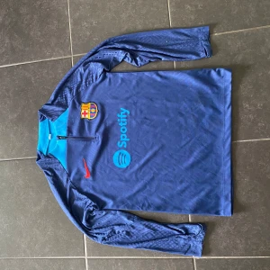 Blå långärmad FC Barcelona träningströja - Säljer en blå långärmad FC Barcelona träningströja från Nike med klubbmärke och röd swoosh på bröstet. Spotify-logga i ljusblått framtill och halv dragkedja vid halsen. Tillverkad i lätt och ventilerande syntetmaterial, perfekt för träning.