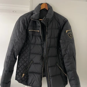 Svart  pufferjacka Rockandblue - Snygg svart pufferjacka från Rockandblue med quiltade partier och guldiga dragkedjor. Jackan har hög krage, flera fickor med dragkedja och en patch på ärmen. Perfekt för kyliga dagar och ger en cool vibe till din outfit.