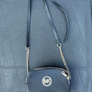 Svart axelväska Michael Kors skinn - Snygg svart axelväska från Michael Kors i skinn med många fack, guldfärgade kedjedetaljer och dragkedjor. Väskan har rundade kanter, MK-logga framtill och justerbar axelrem i både läder och kedja. Perfekt storlek för det viktigaste och stilren look.